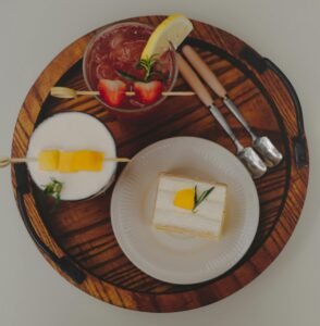 Aesthetic flatlay featuring a cake slice, fruity cocktails, and cutlery on a rustic wooden tray.