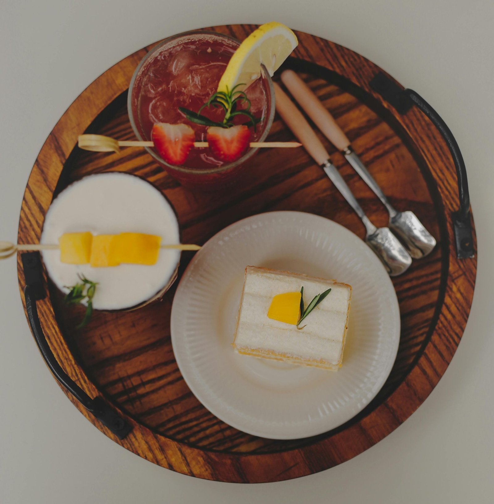 Aesthetic flatlay featuring a cake slice, fruity cocktails, and cutlery on a rustic wooden tray.